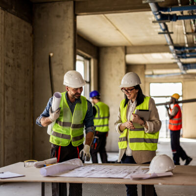 Male and female architects reviewing renovation blueprints with builders working in the background.